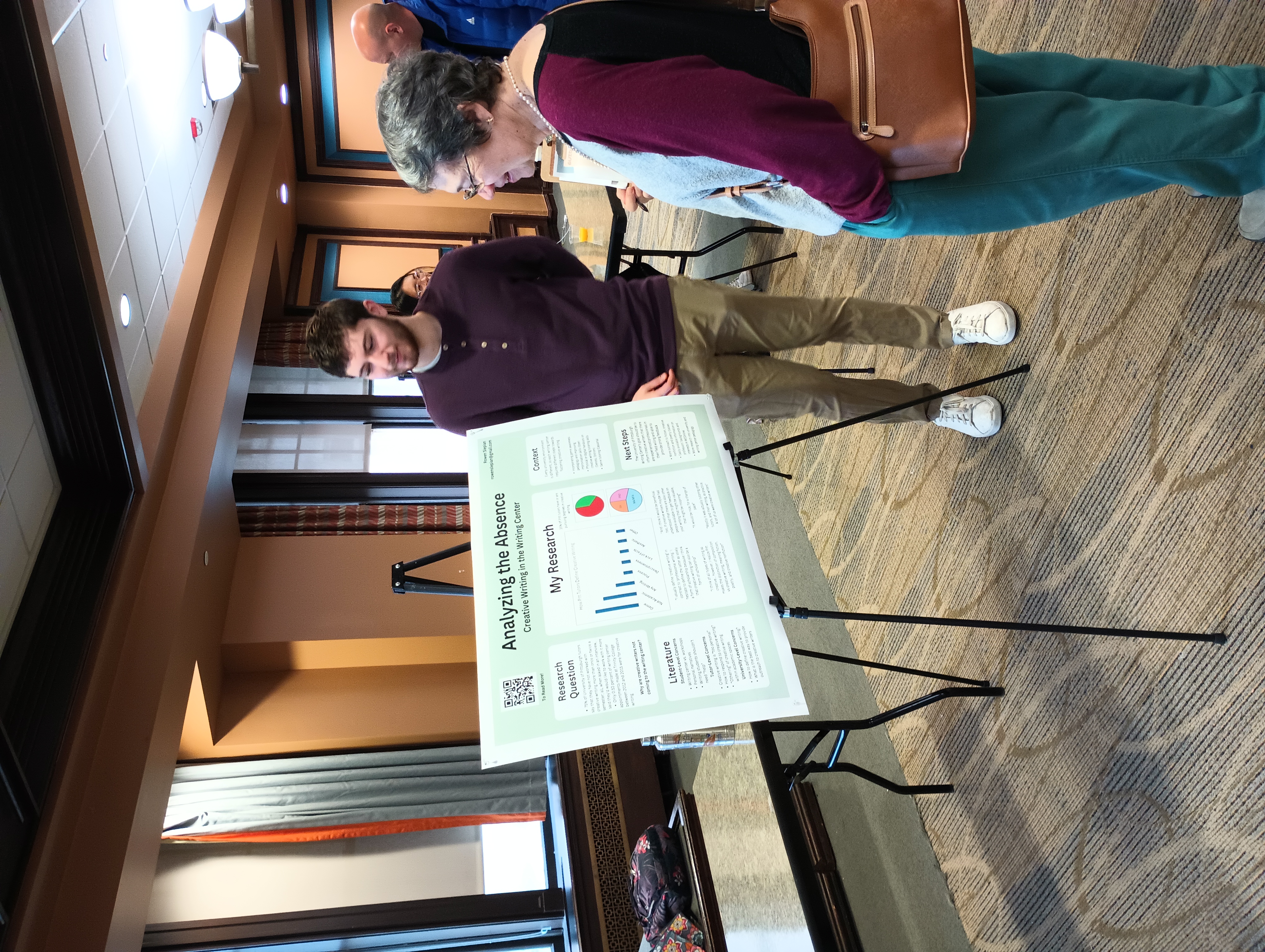 A peer tutor presents their poster to a faculty member. The poster is on a tripod. The poster title is legible and reads "Analyzing the Absence."