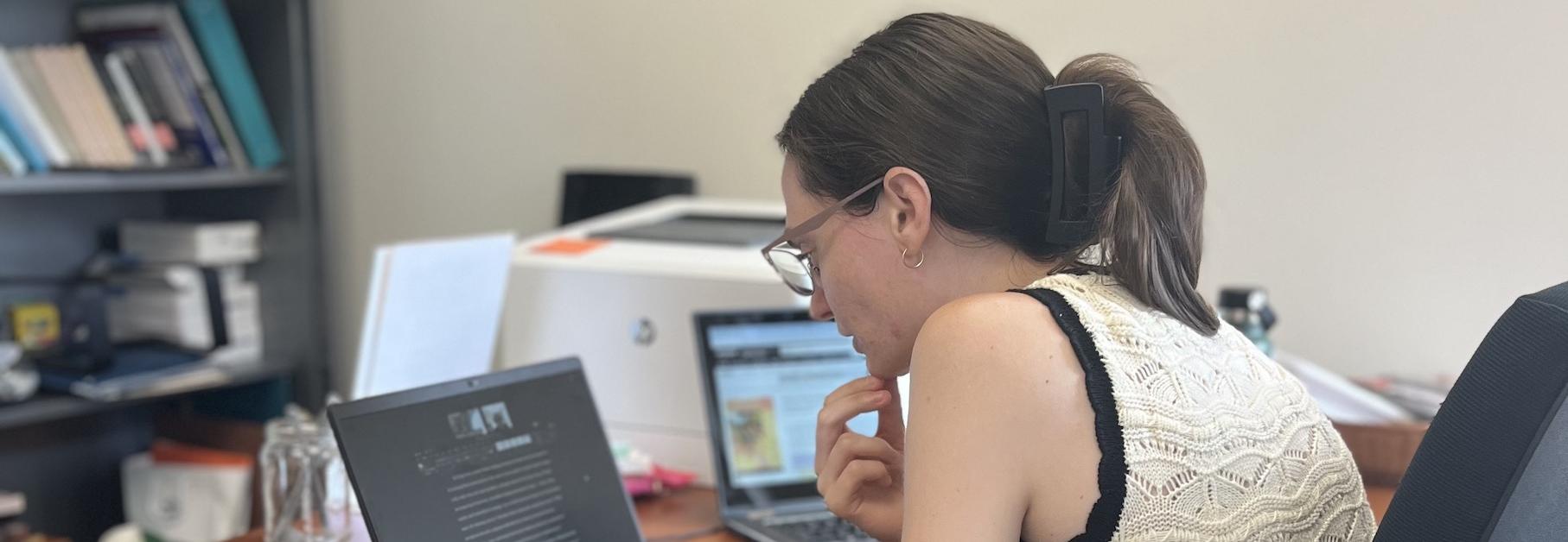 A writing tutor reads a document and works with a student over a laptop in an office.