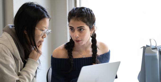 A student and a peer tutor in discussion with a laptop screen between them.  