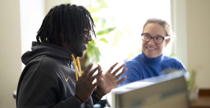 A student and faculty consultant work together seated at a desk. The student has their hands open and is smiling. The faculty consultant smiles at the student.