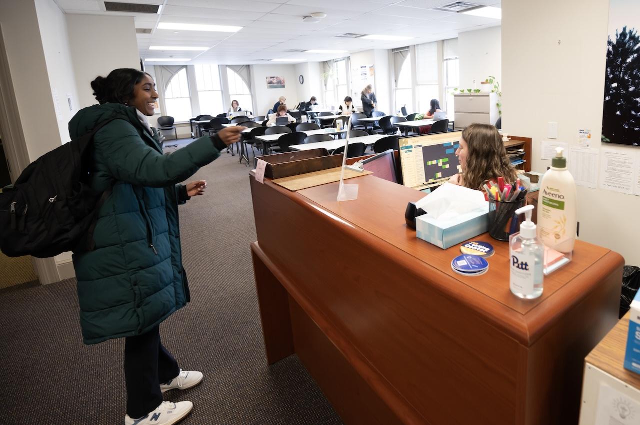 Student turning in form to office manager who is sitting behind a large desk. 